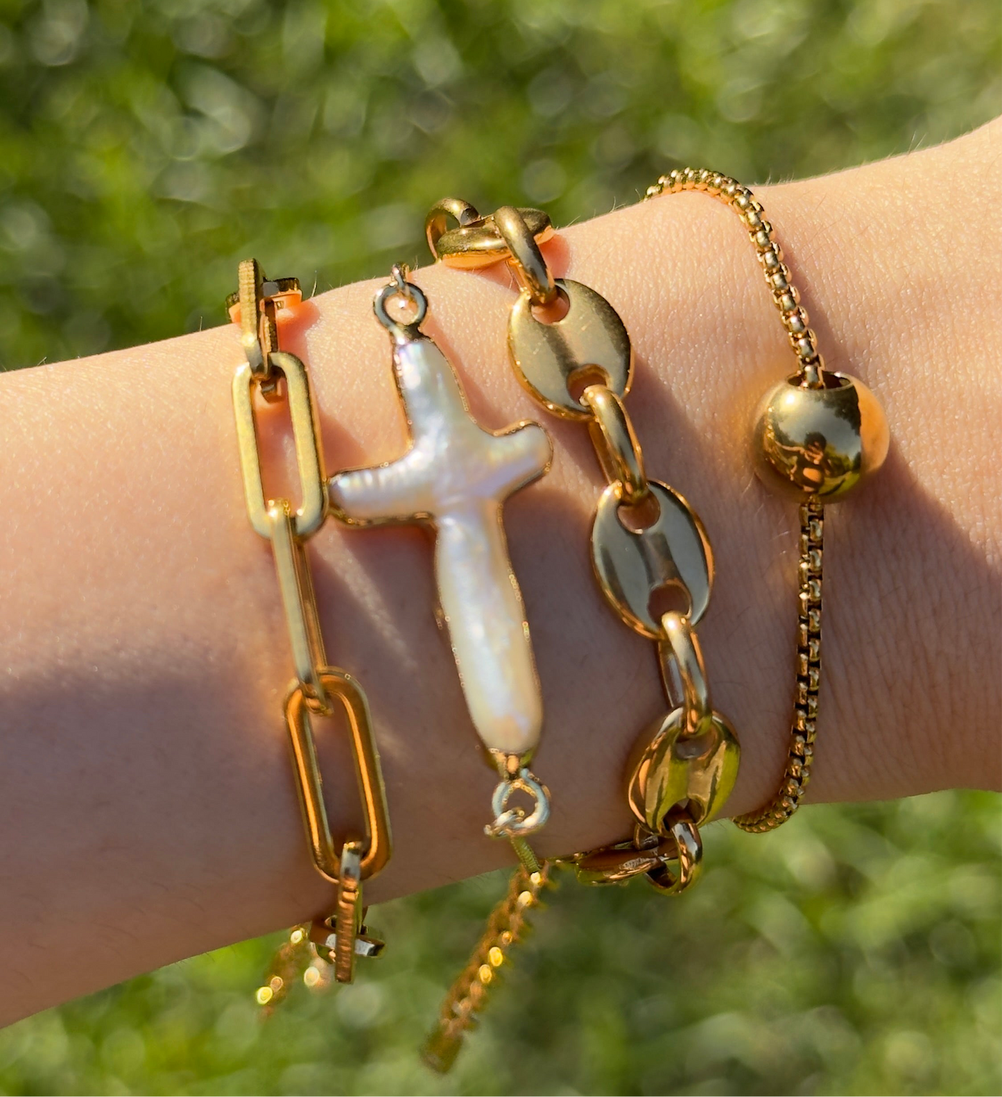 Gold bracelets with a cross charm on a wrist against a blurred green background