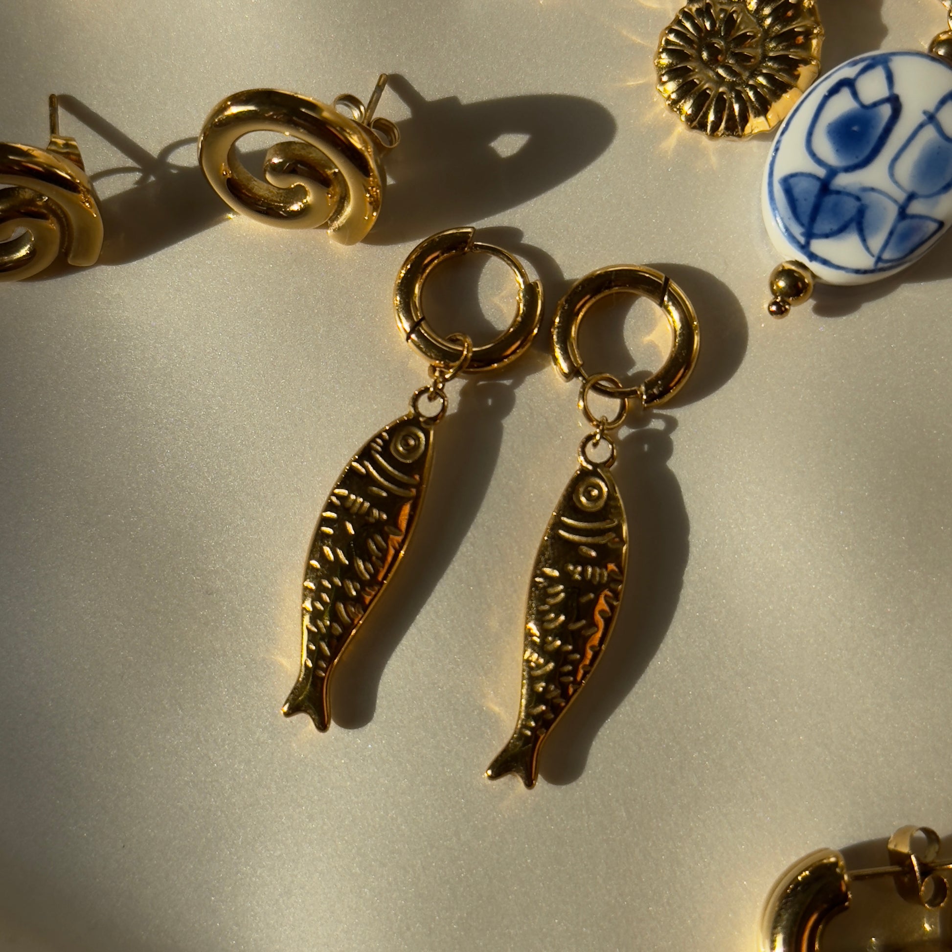 Gold fish-shaped earrings on a light gray surface with other jewelry items.