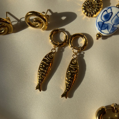 Gold fish-shaped earrings on a light gray surface with other jewelry items.